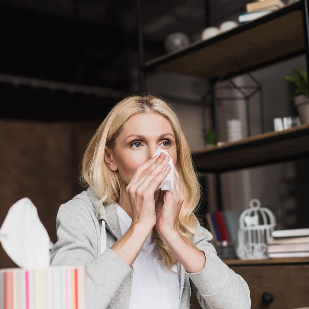 sick middle aged woman with runny nose holding paper towel and looking away at home